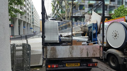A flatbed truck parked on the side of a city street, loaded with various discarded household items and rubbish, including white wooden panels, a grey metal furniture frame, and some packaging materials. The truck is positioned near a modern residential building with several balconies visible in the background. The vehicle's cargo area appears cluttered with mixed waste suitable for household rubbish removal services in Borehamwood. The scene is illuminated by natural daylight, with the urban environment surrounding the vehicle indicating a typical house clearance or domestic waste collection context. The truck's licence plate reads MX12 GHA, and it is part of a rubbish removal operation perhaps conducted by House Clearance Borehamwood, specializing in clearance services including home, loft, and furniture removal in the local area.