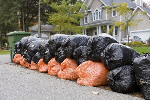 A row of black and orange plastic rubbish bags filled with waste material, neatly stacked along the curbside on a residential street. The black bags are on top, with some appearing slightly shiny and tightly packed, while the orange bags are positioned underneath and are somewhat less reflective, indicating a softer plastic. The setting includes a paved pavement and an asphalt road, with a background of suburban houses with white and grey facades, some with porches and gabled roofs. Green trees with fresh leaves line the street, suggesting spring or early summer. The scene is illuminated by natural daylight, creating a clear and neutral atmosphere suitable for a rubbish removal service based in Borehamwood, where House Clearance Borehamwood offers professional waste collection and clearance in the local area.