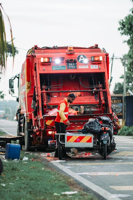 A pile of assorted waste materials and rubbish is gathered on a paved outdoor area, with black plastic rubbish bags, cardboard boxes, and discarded items such as an old, worn upholstery armchair with 
