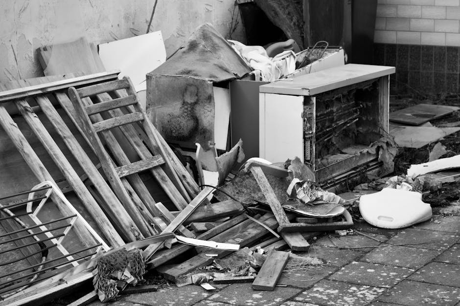 A collection of discarded household items and debris arranged on a paved outdoor surface against a brick and wooden wall background. Prominently, there are two wooden bedsprings lacking mattress, lean