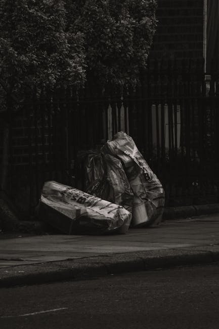 A collection of discarded household items and debris arranged on a paved outdoor surface against a brick and wooden wall background. Prominently, there are two wooden bedsprings lacking mattress, lean