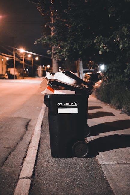 A black wheeled rubbish bin labeled 'ST. JOHN'S' positioned on a sidewalk at night, with its lid open revealing an overflowing collection of mixed waste materials, including crumpled paper, plastic ba